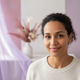 A smiling woman in her late 30s, representing a happy, trusted customer. The background is softly blurred with dusty rose and